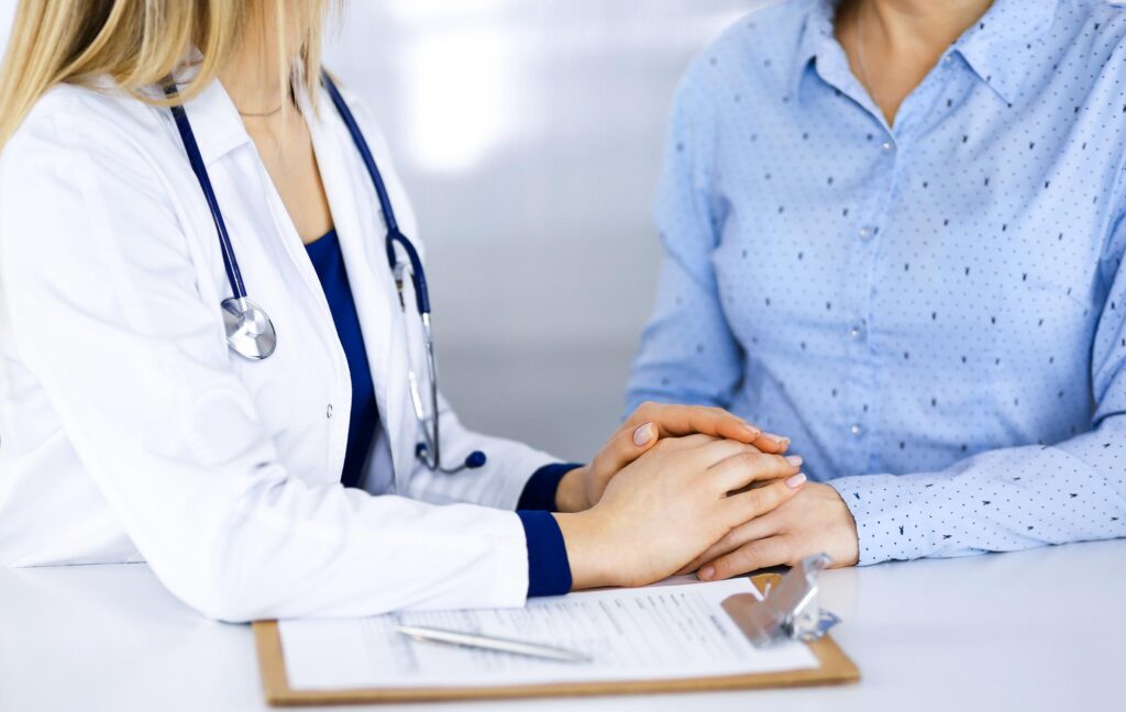 Woman Doctor with Woman Patient talking at a desk, doctor holding patient's hand - Dr Tracy Pfeifer - New York City and The Hamptons